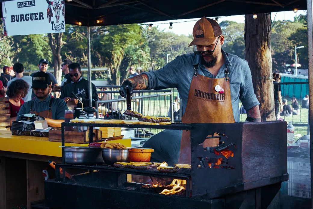 Ein Koch bereitet Burger auf einem offenen Grill an einem Essensstand im Freien zu; im Hintergrund stehen Gäste