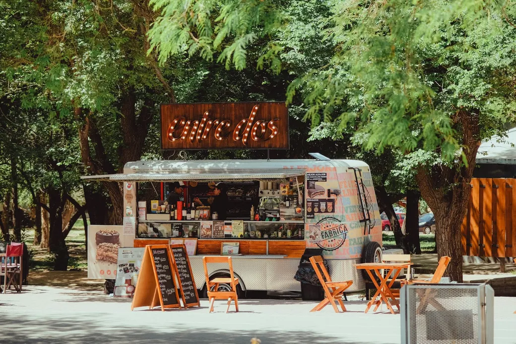 Foodtruck unter grünen Bäumen mit geöffneter Verkaufsklappe, Tischen und orangefarbenen Stühlen davor